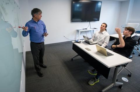 two people sitting at a table with laptops and one person standing by a whiteboard in the UNH Entrepreneurship Center